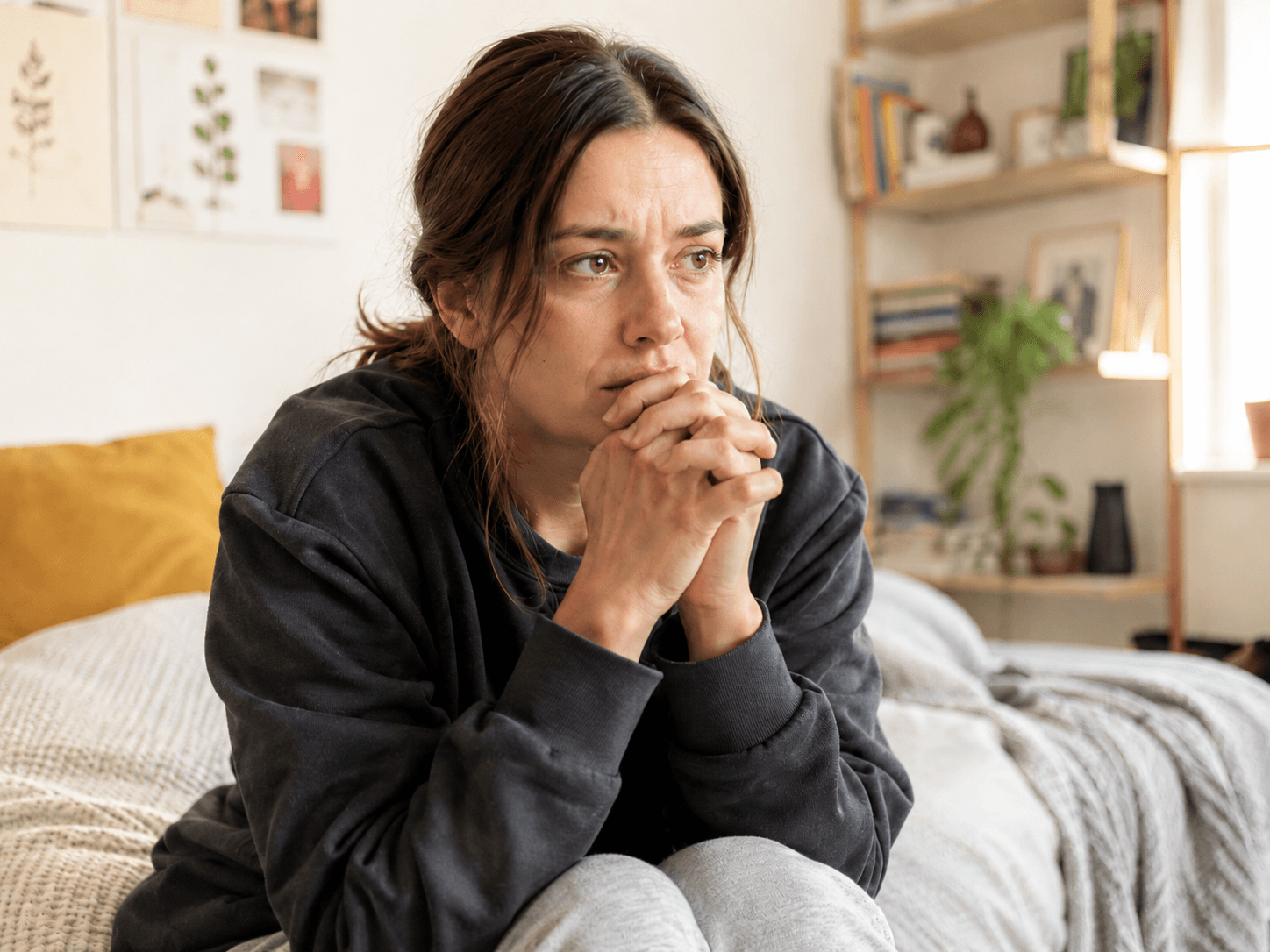 A woman sitting on a bed with clasped hands and a worried expression.