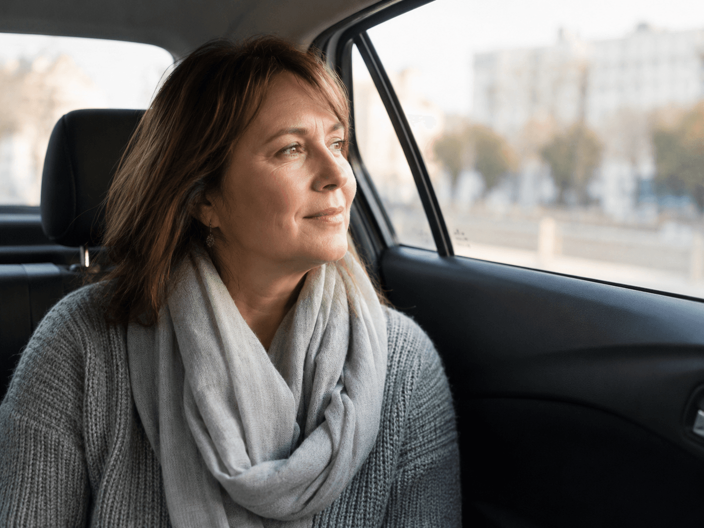 A woman with a scarf looking out the window from the back seat of a car.